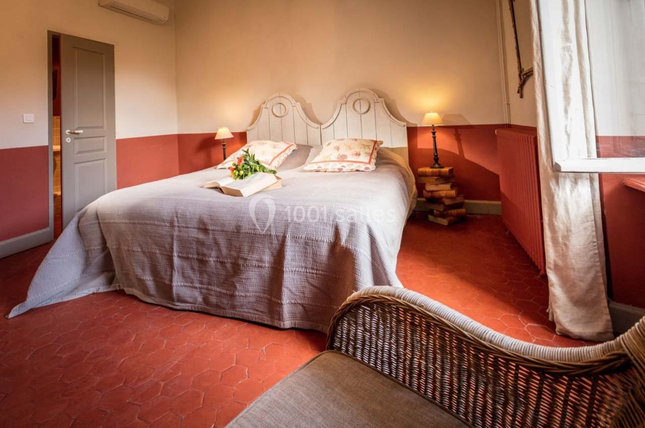 Chambre avec un grand lit, couvre-lit gris, coussins fleuris, lumière naturelle et sol en tomettes rouges.