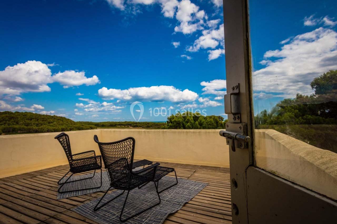 Deux chaises noires sur une terrasse en bois avec vue sur un paysage verdoyant sous un ciel bleu parsemé de nuages.