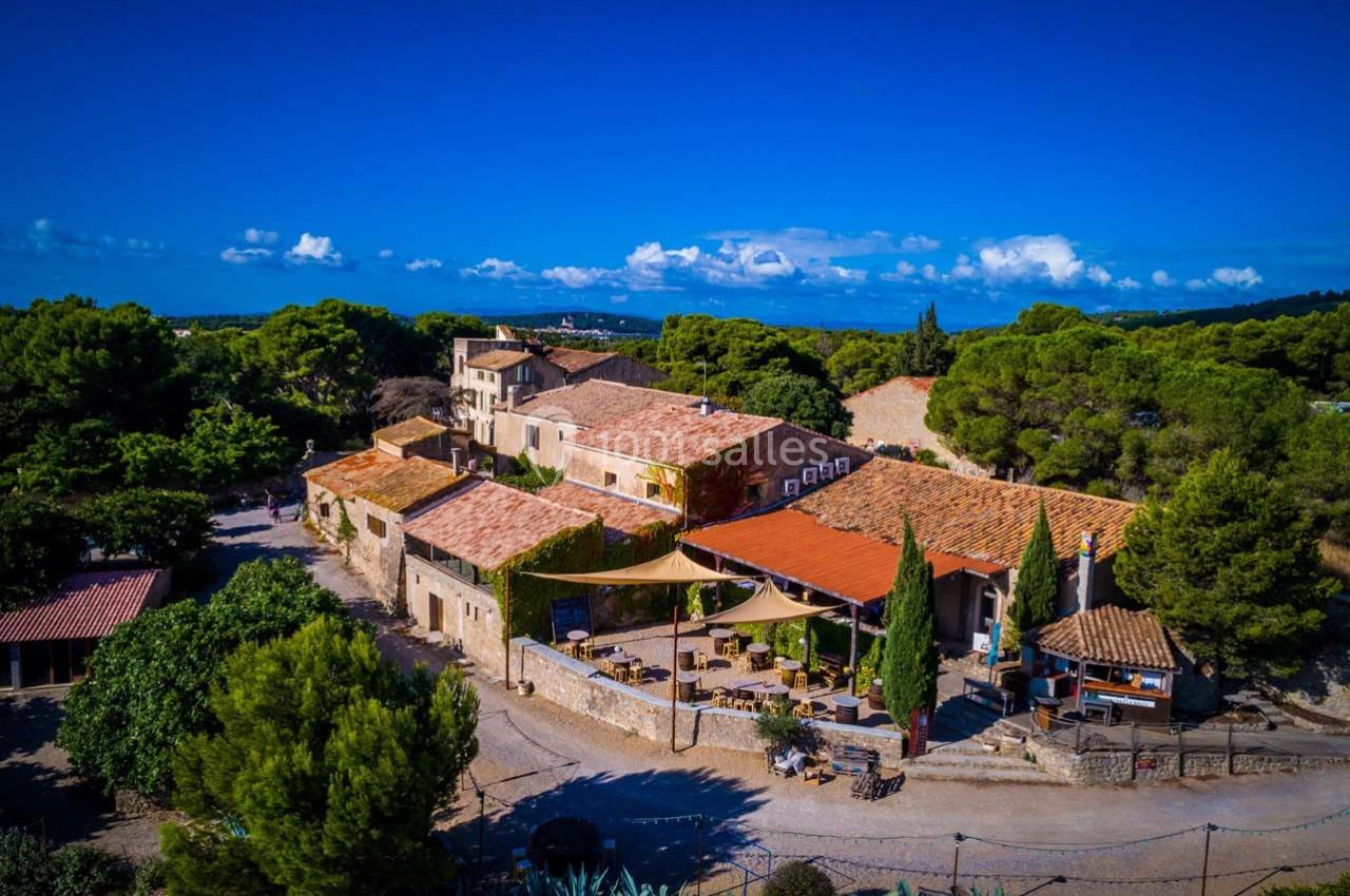Vue aérienne d'un domaine en pierre avec terrasses ombragées, entouré de végétation et sous un ciel bleu dégagé.