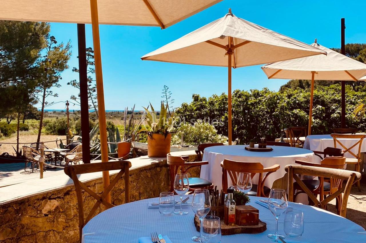 Terrasse ensoleillée d'un restaurant avec tables dressées, chaises en bois et parasols, entourée de verdure.