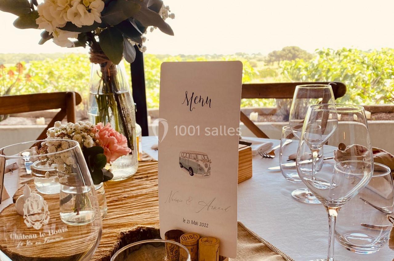 Table élégamment dressée avec fleurs, verres et menu, vue sur un paysage extérieur ensoleillé à travers une baie vitrée.