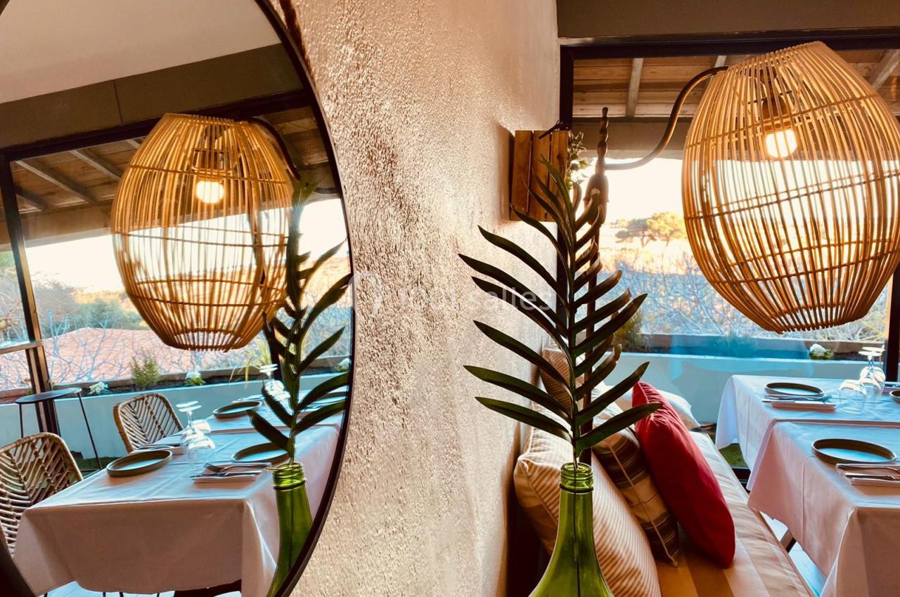 Salle de restaurant lumineuse avec tables dressées, reflet dans un miroir et suspension en osier.