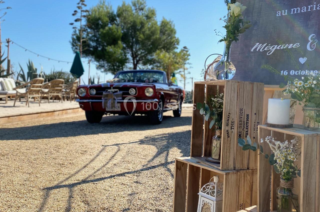 Voiture ancienne rouge garée près d'une décoration de mariage en bois avec fleurs et lanternes, en extérieur ensoleillé.