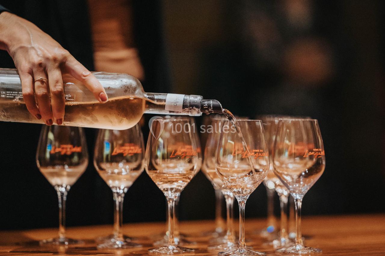 Une main verse du vin blanc dans des verres alignés sur une table en bois.