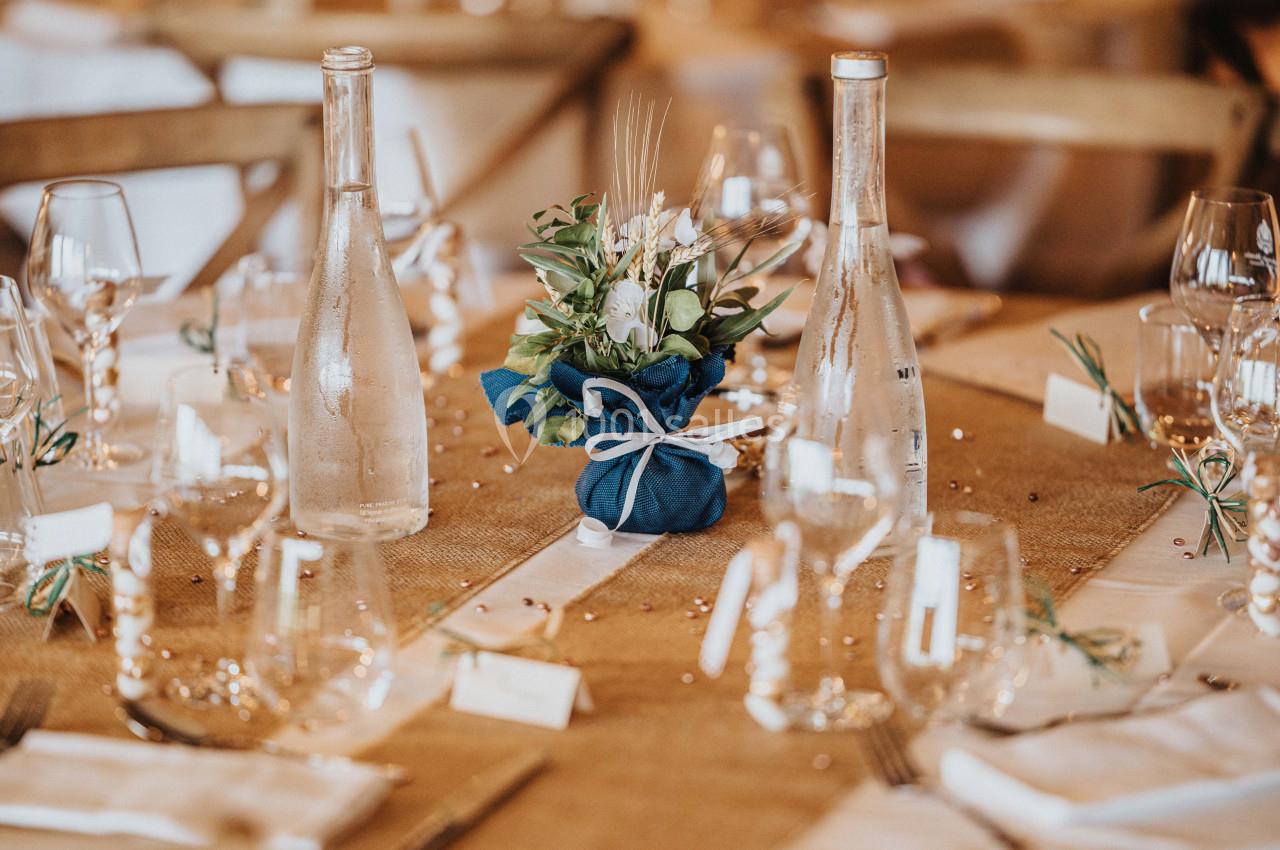 Table décorée avec nappes en jute, bouteilles en verre, verres, et un centre de table floral dans un tissu bleu.