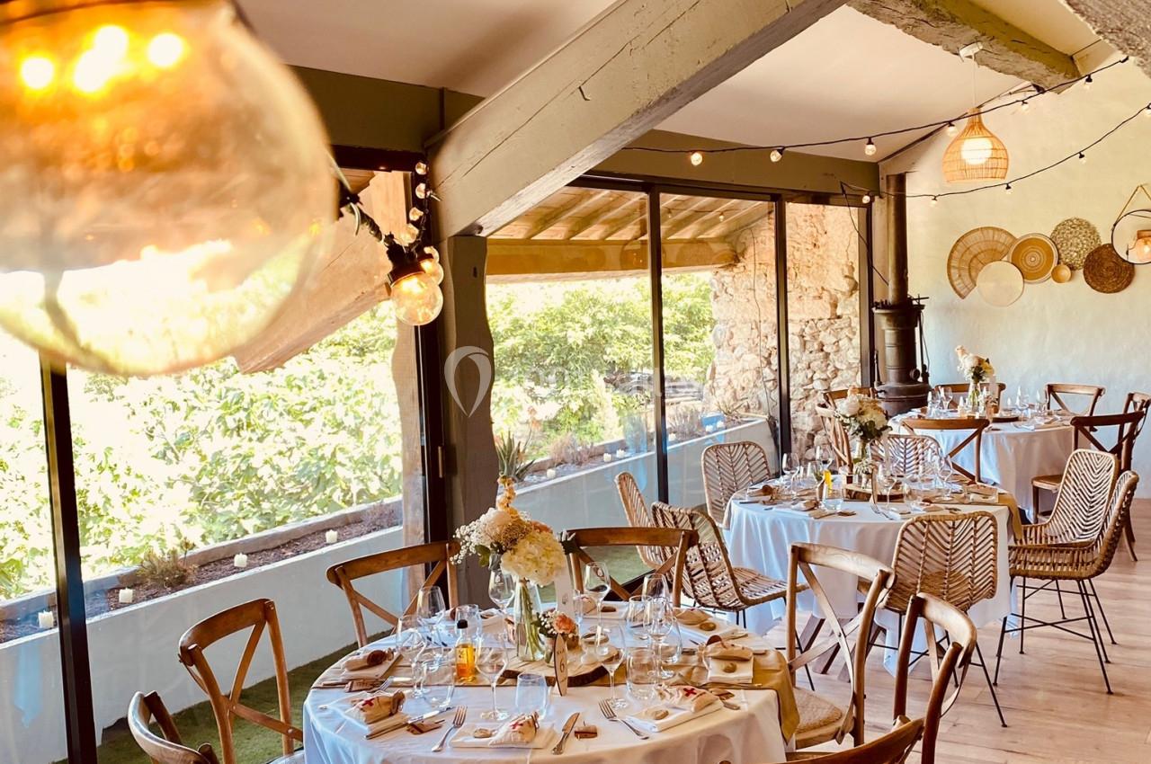Salle de réception lumineuse avec tables rondes dressées, chaises en bois et vue sur un extérieur verdoyant.