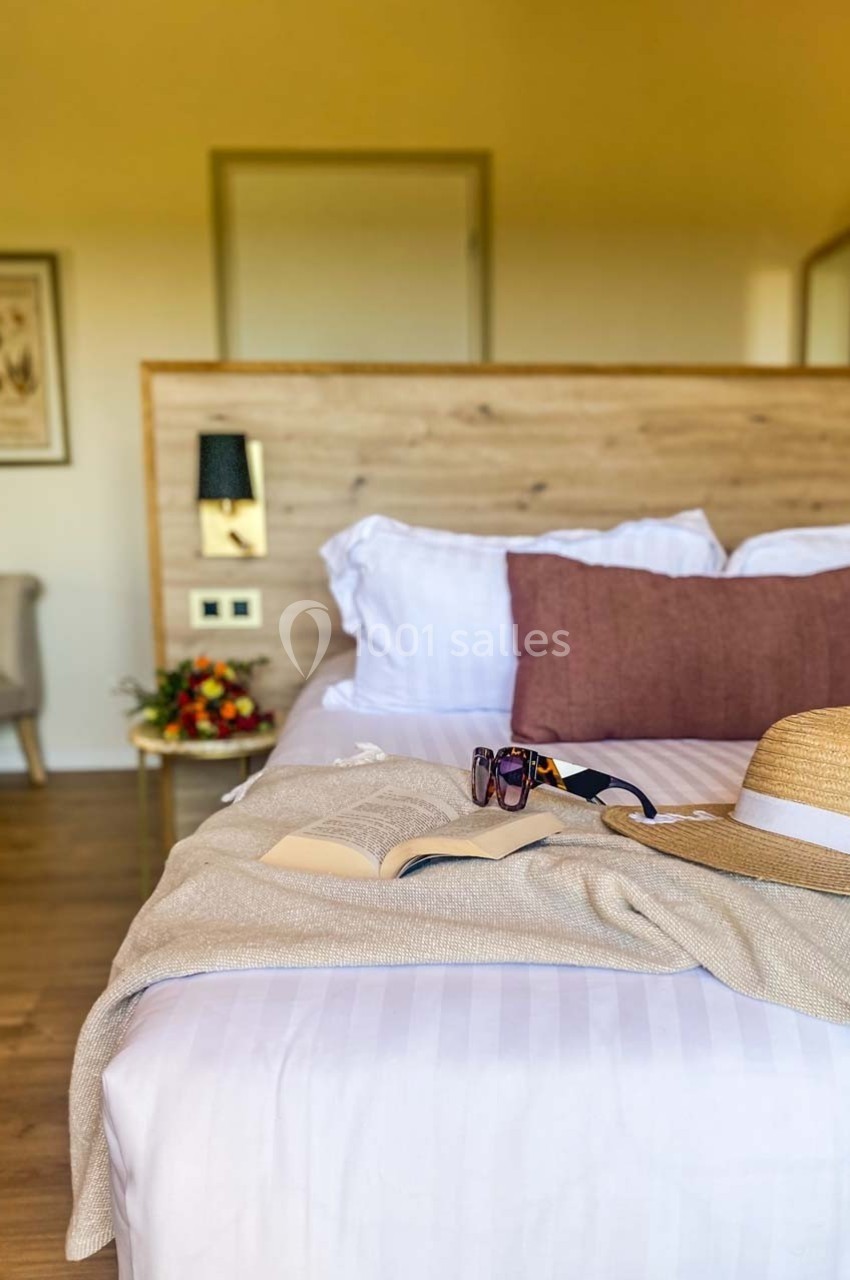 Lit double avec coussins, chapeau, lunettes de soleil et livre posé dessus, dans une chambre lumineuse au décor chaleureux.