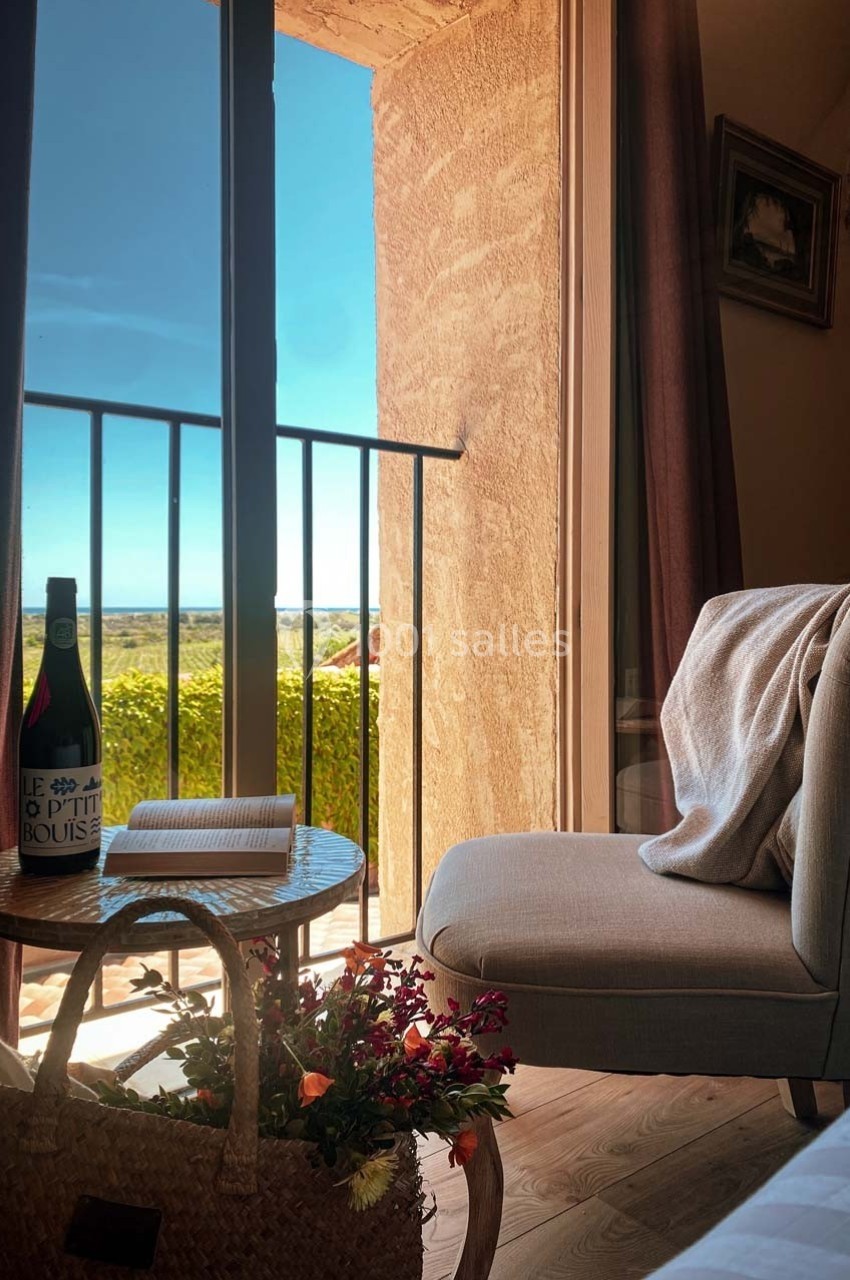 Fauteuil près d'une table avec un livre, une bouteille de vin et un panier de fleurs, face à une fenêtre ouverte sur un…