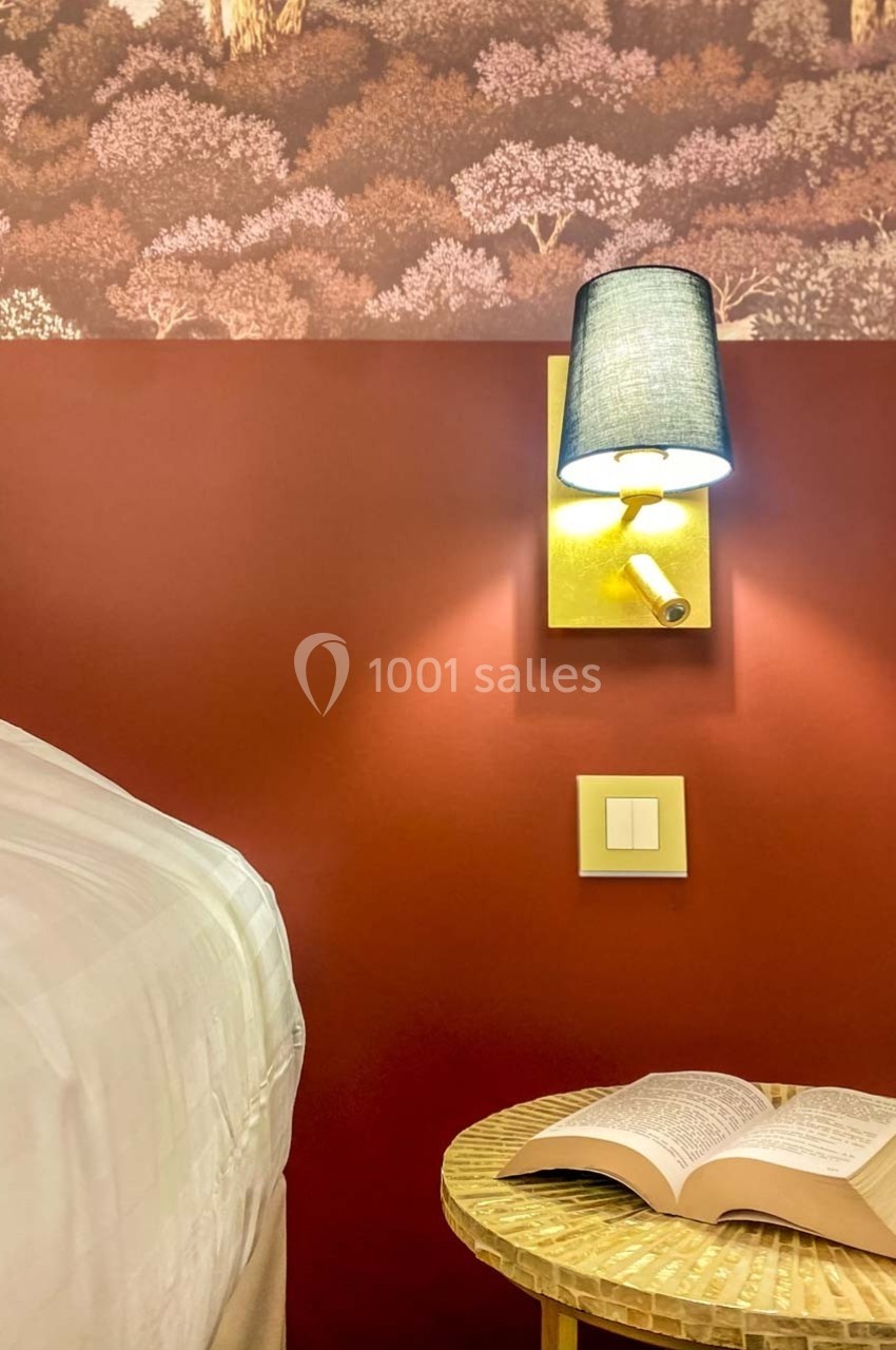 Lampe murale allumée au-dessus d'une table en rotin avec un livre ouvert, près d'un lit et un mur rouge décoré.