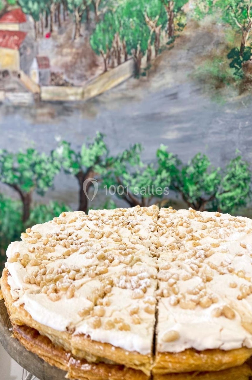 Tarte garnie de crème et saupoudrée de pignons de pin, posée devant une fresque représentant un paysage rural.