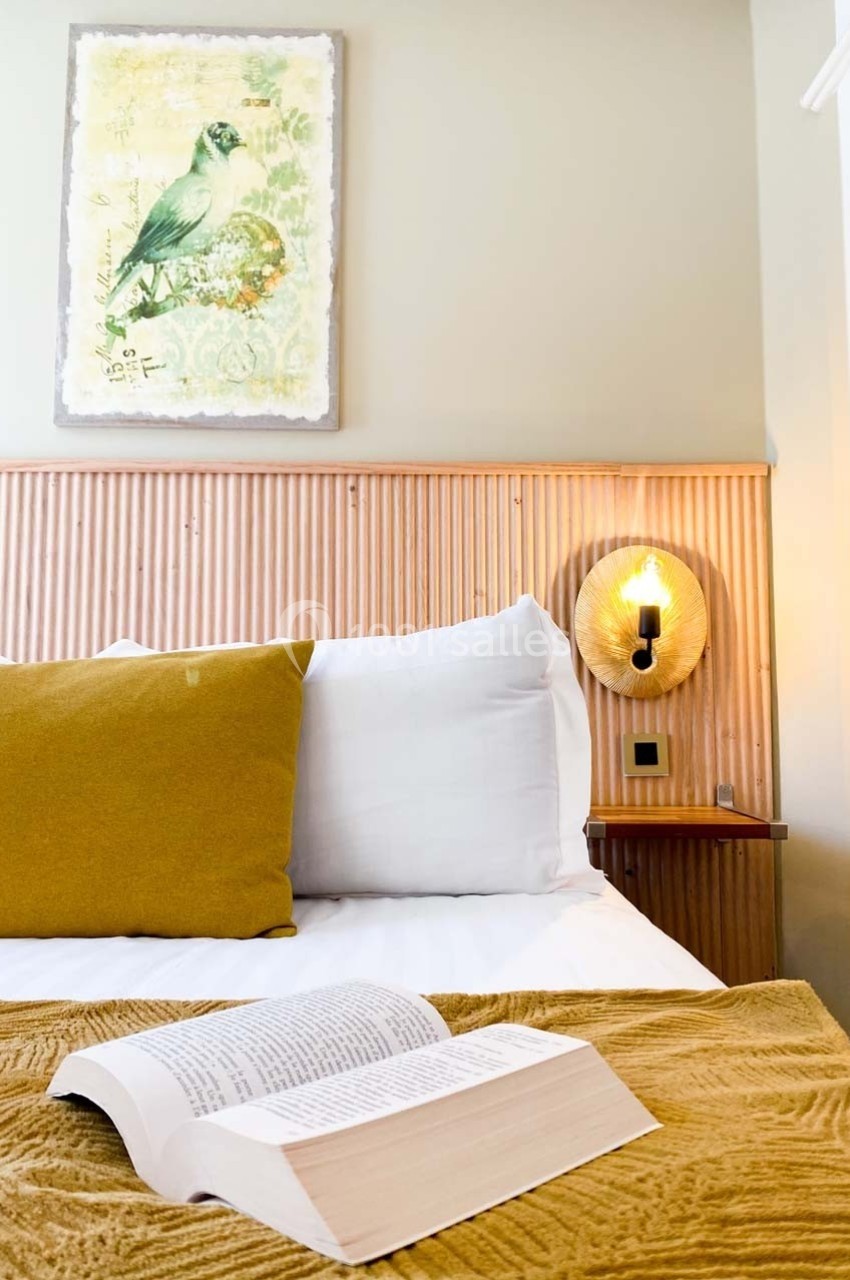 Lit avec coussins jaunes, couverture assortie, livre ouvert au premier plan et lampe murale allumée en arrière-plan.