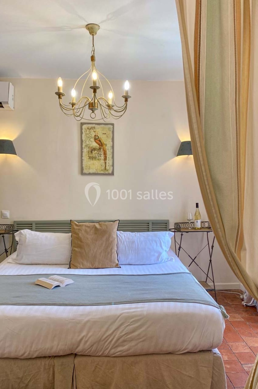 Lit double avec coussins, draps clairs, tables de chevet, rideaux ouverts, lustre et tableau mural dans une chambre…