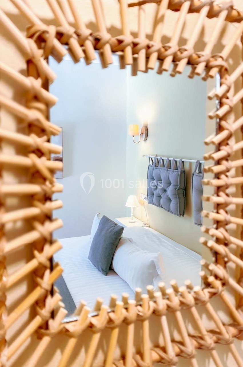 Reflet d'une chambre lumineuse avec lit double, coussins gris et lampe de chevet, dans un miroir à cadre en rotin.