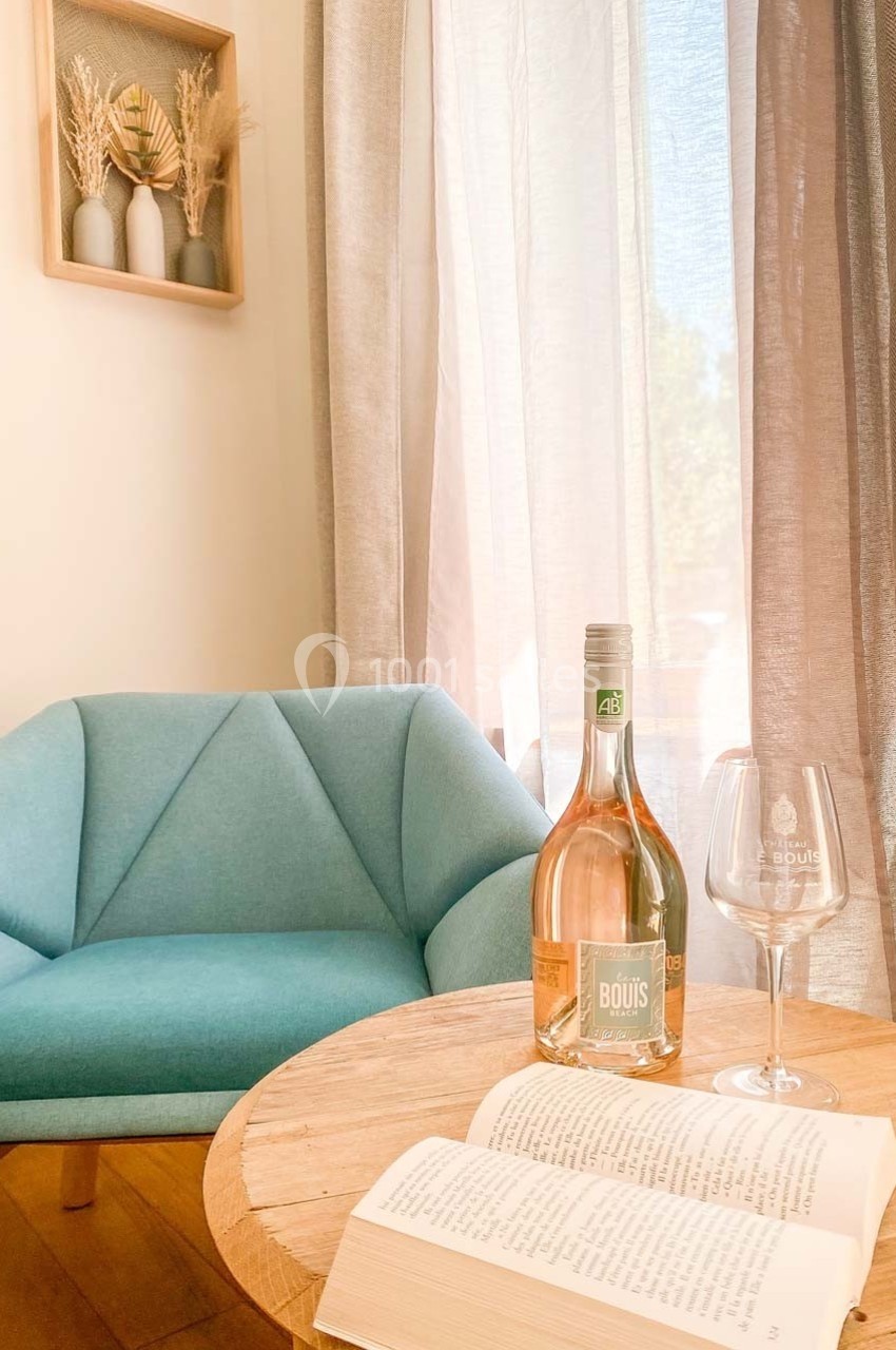 Une bouteille de vin rosé, un verre, un livre ouvert sur une table ronde près d'un fauteuil bleu et d'une fenêtre.