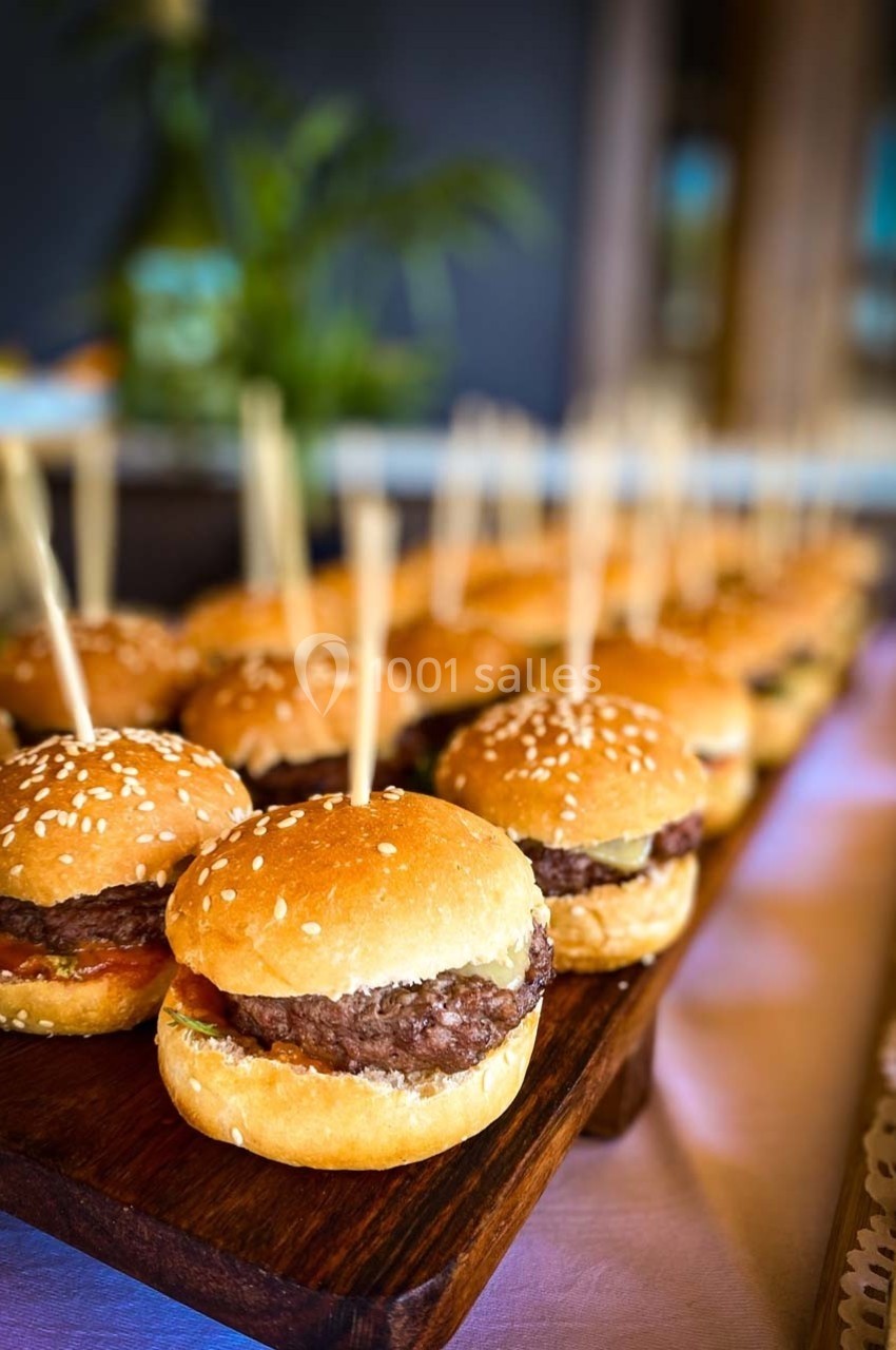 Plateau de mini-burgers avec pain aux graines de sésame, garnis de viande et de condiments, présentés sur une table en bois.
