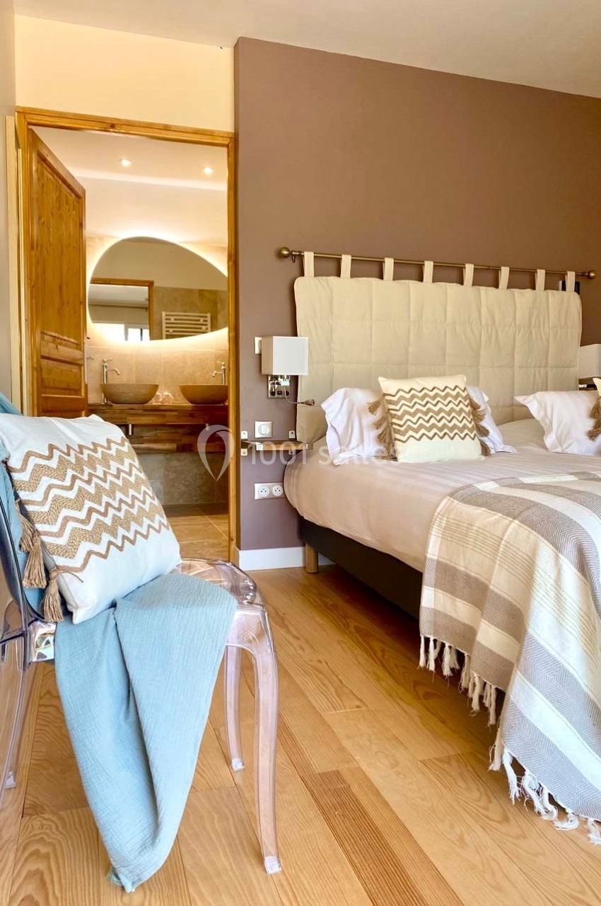 Chambre lumineuse avec lit double, plaid et coussins, vue sur une salle de bain ouverte avec lavabo et miroir.