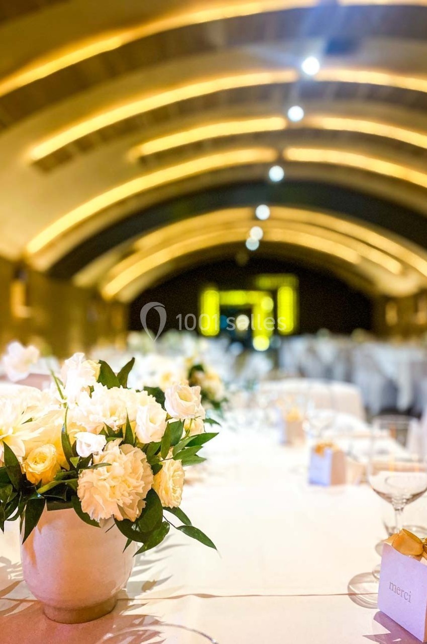 Salle voûtée décorée pour un événement, avec des tables dressées et un bouquet de fleurs blanches au premier plan.
