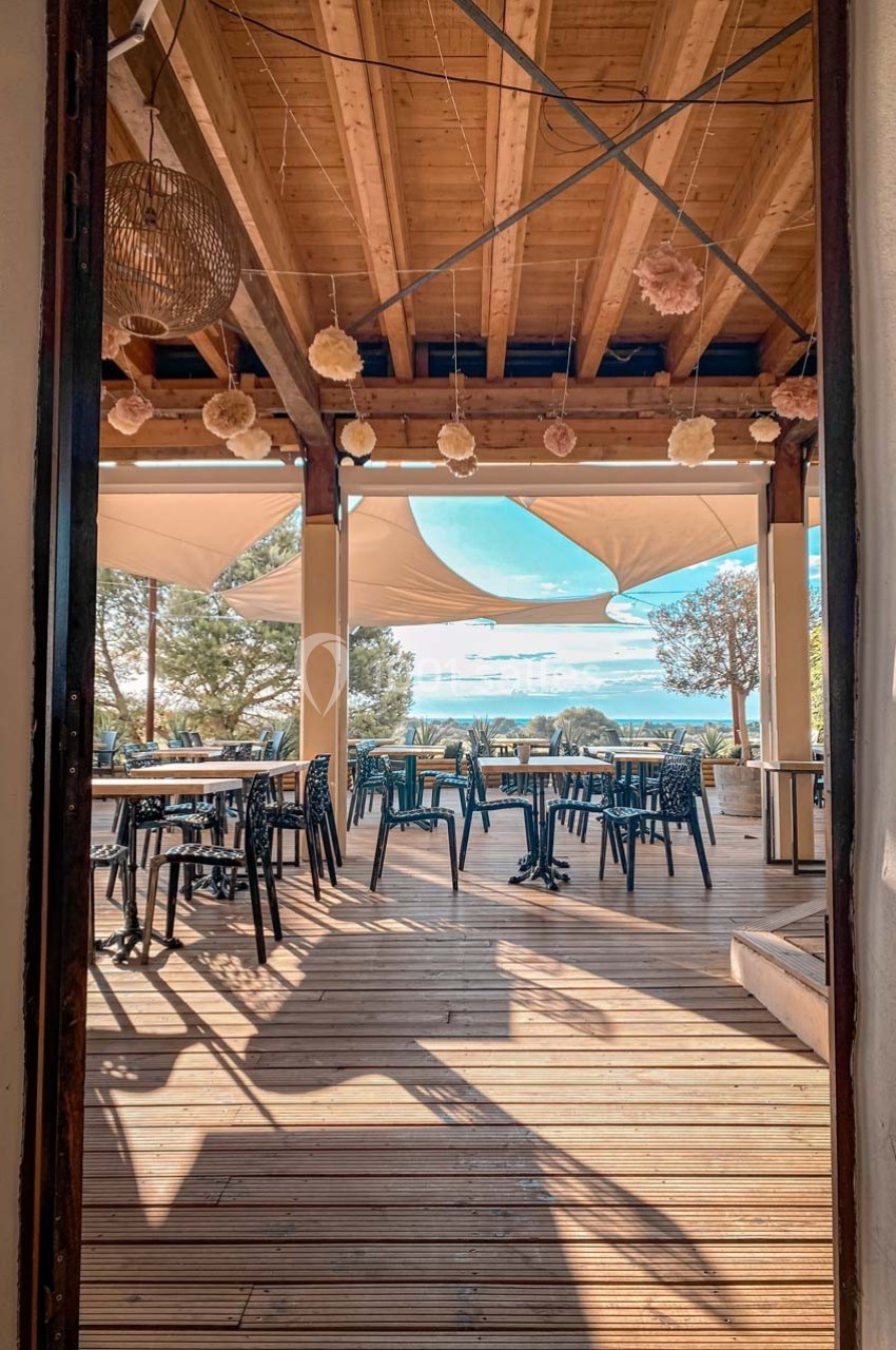 Terrasse en bois avec tables et chaises, ombragée par des voiles tendues, offrant une vue dégagée sur un paysage ensoleillé.