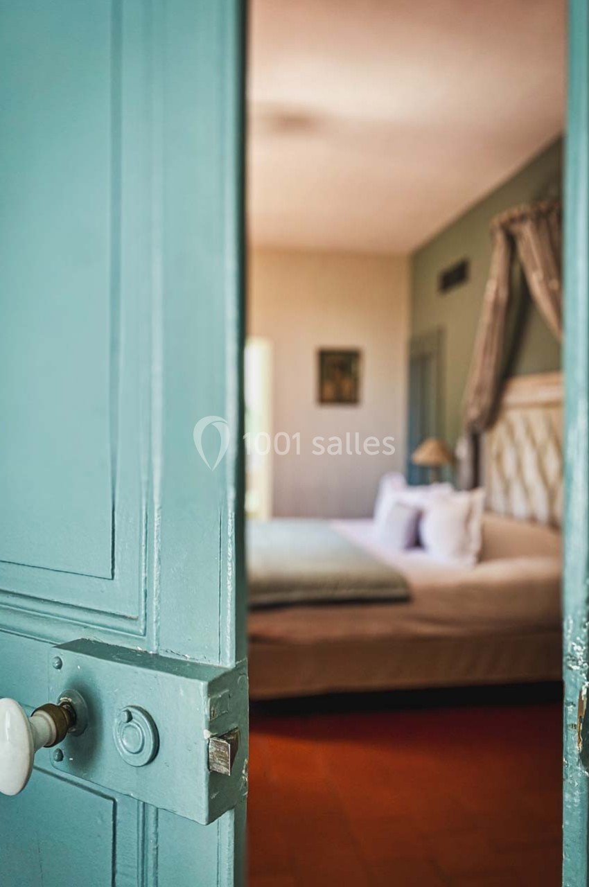 Porte bleue entrouverte donnant sur une chambre lumineuse avec un lit double et des draperies.