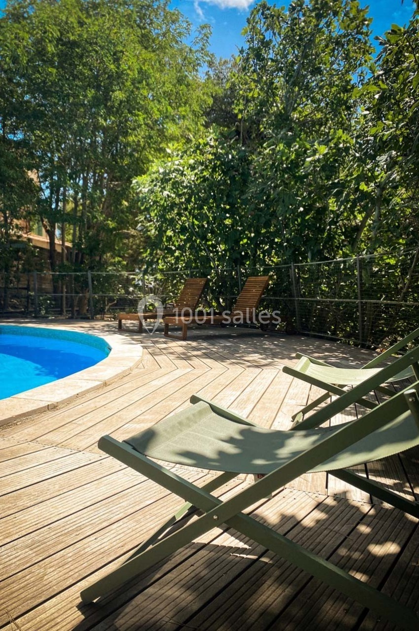 Chaises longues vertes sur une terrasse en bois près d'une piscine, entourée de végétation dense sous un ciel bleu.