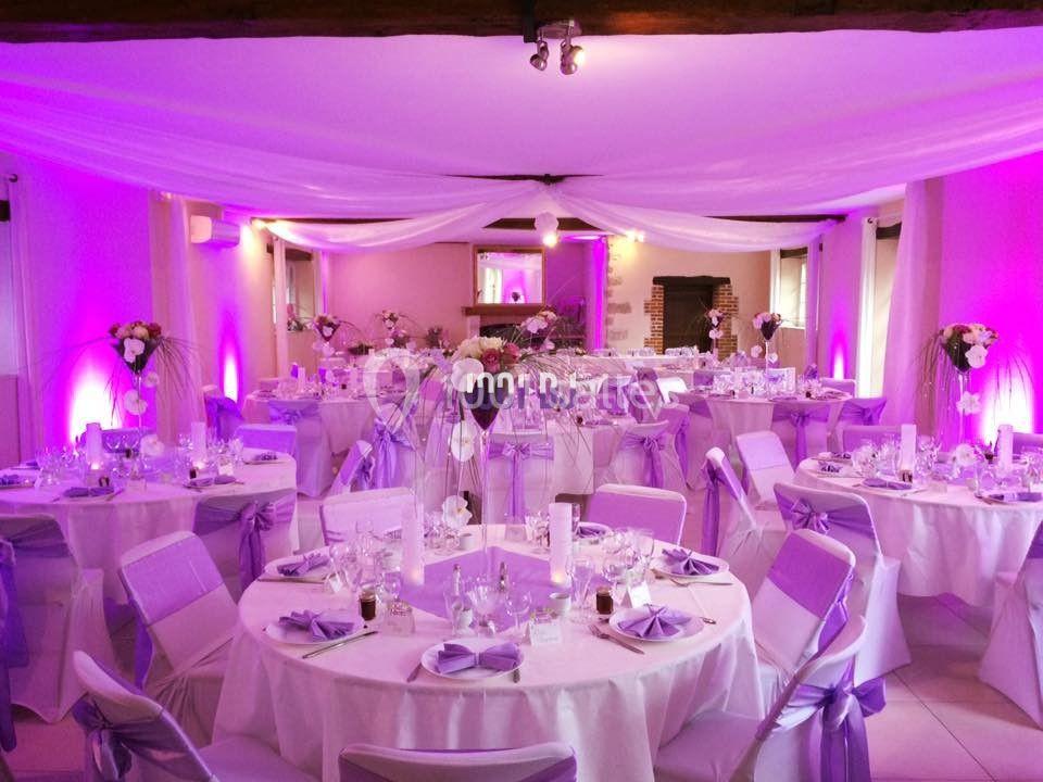 Salle de réception décorée avec des nappes et des rubans violets, éclairage tamisé et tables dressées.