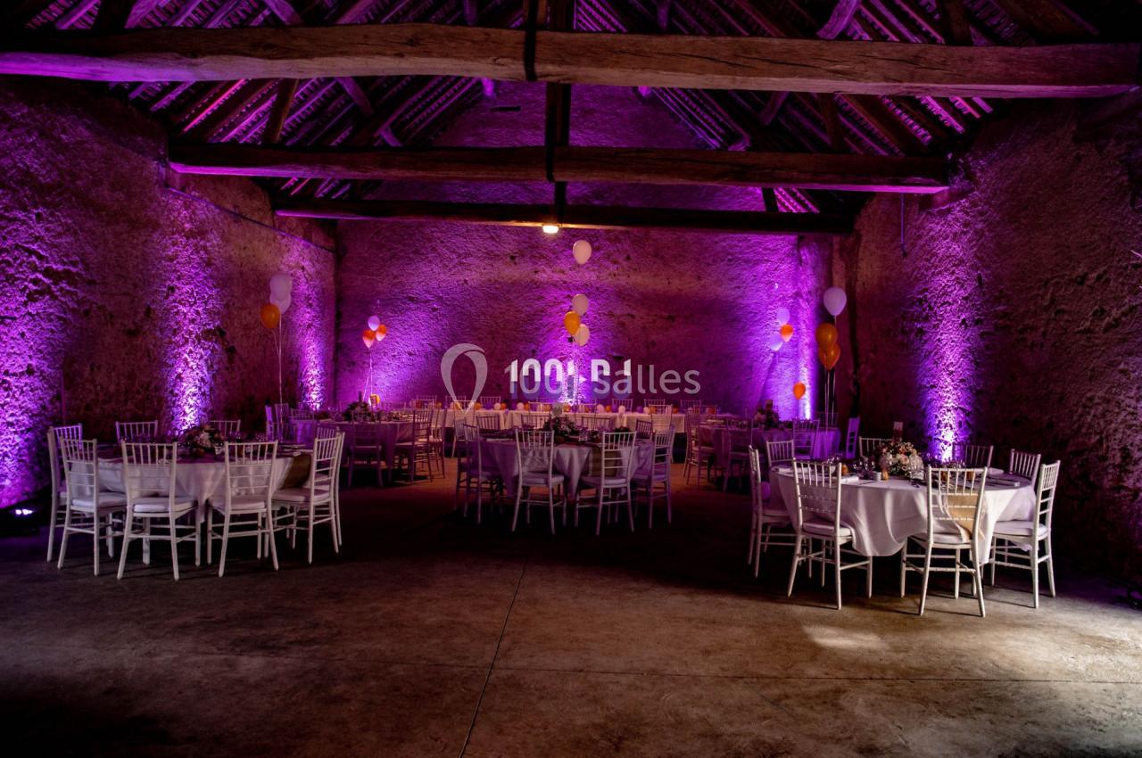 Salle de réception décorée avec des tables rondes, des chaises blanches et un éclairage violet sur les murs en pierre.