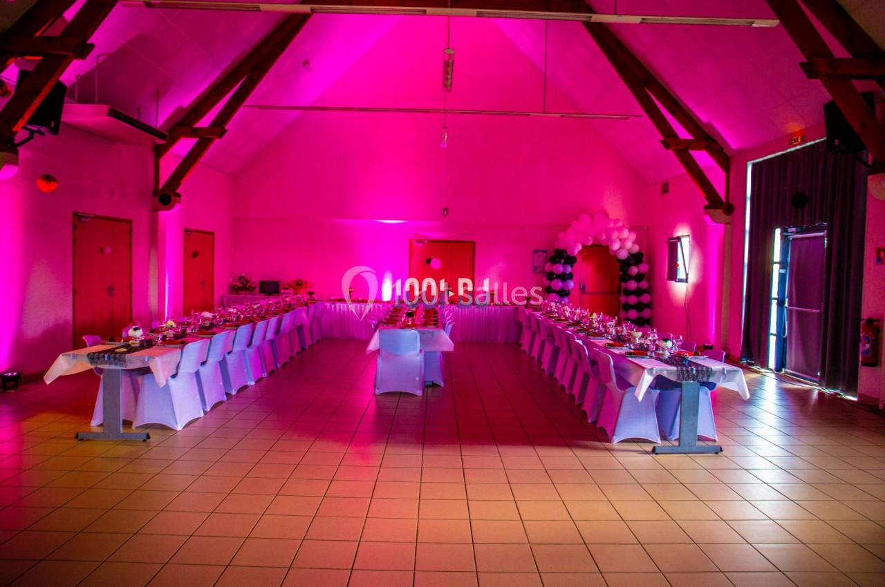 Salle de réception décorée avec des tables alignées, éclairage rose et arche de ballons en arrière-plan.