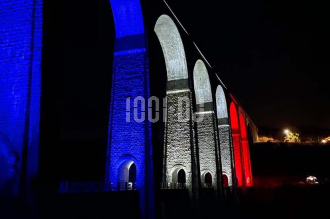 Viaduc illuminé aux couleurs bleu, blanc et rouge dans un paysage nocturne.
