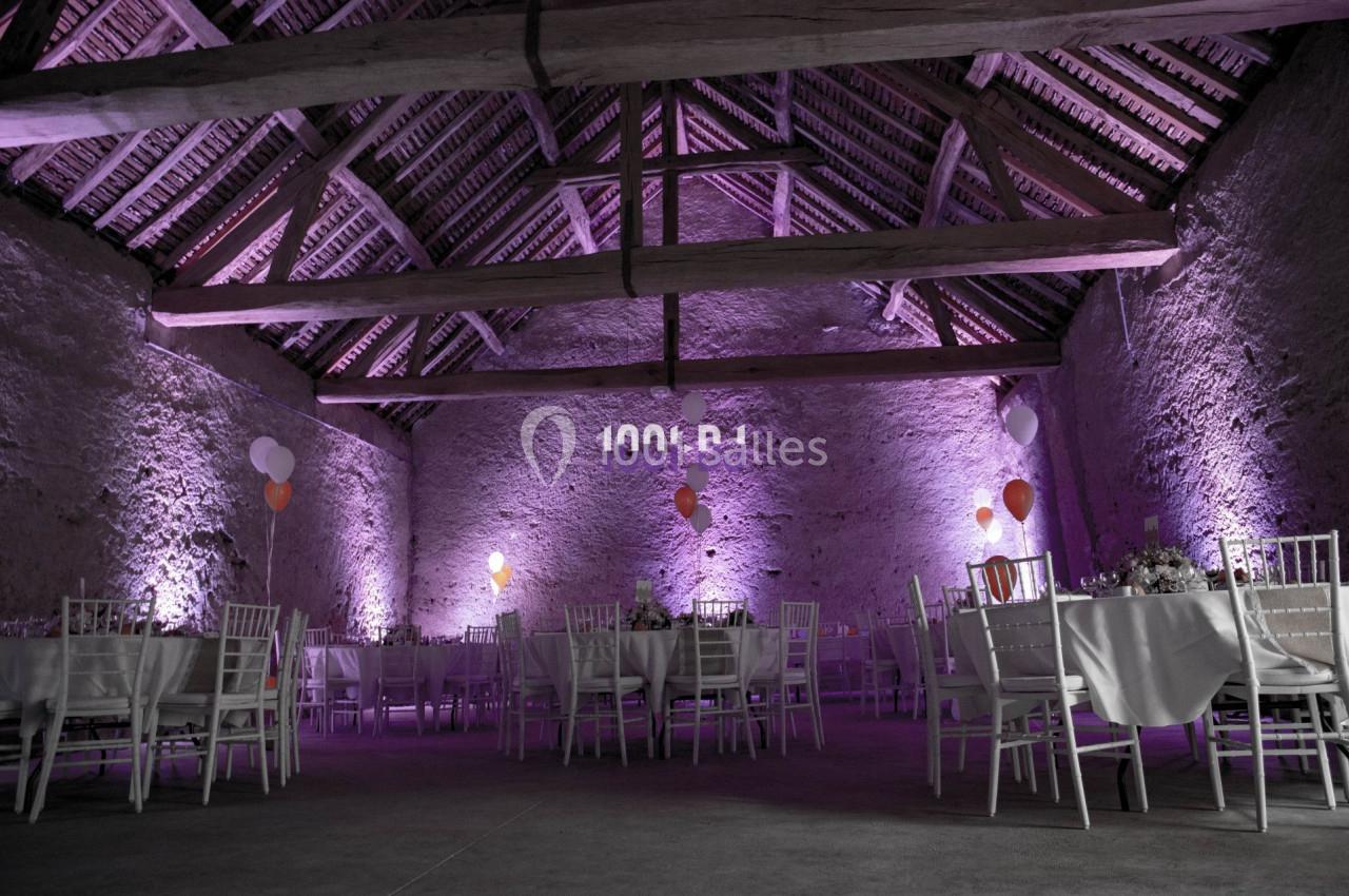 Salle en pierre avec charpente apparente, éclairée en violet, décorée de ballons et tables dressées pour un événement.