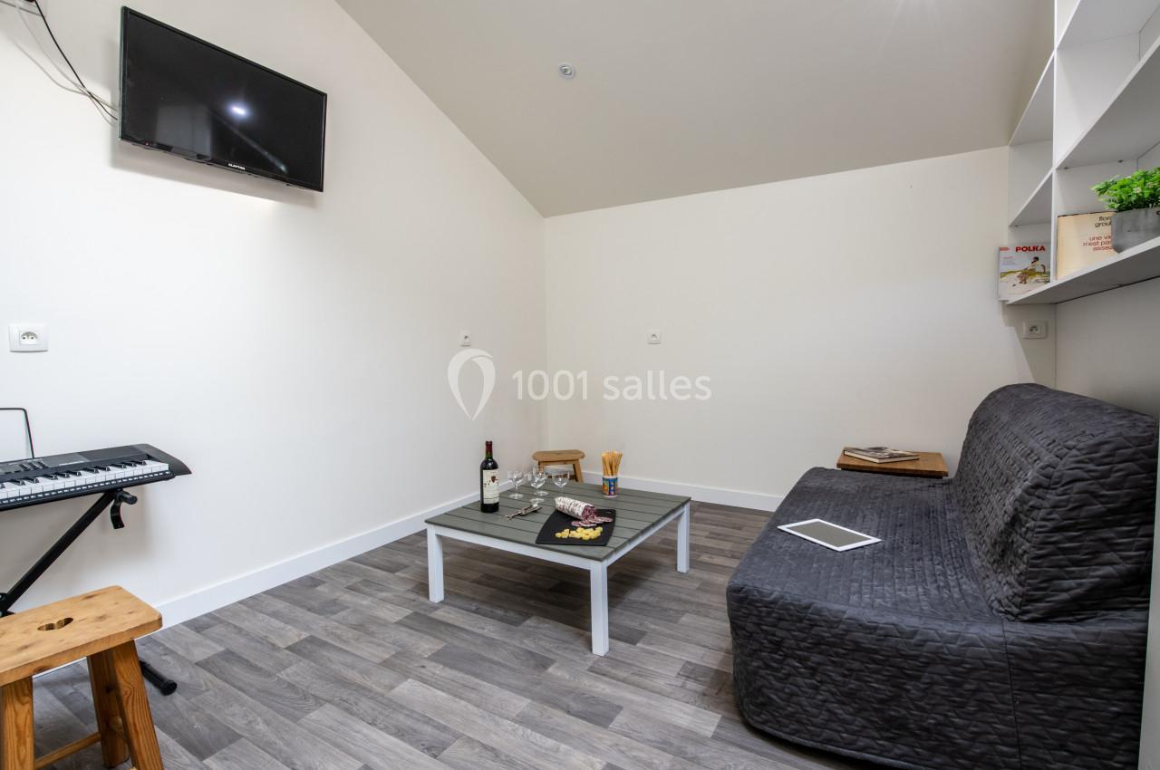 Salon avec canapé gris, table basse avec snacks et vin, étagères murales, clavier musical et télévision fixée au mur.