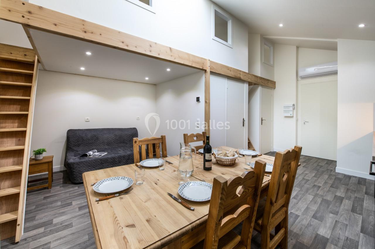 Salle à manger avec table en bois dressée, canapé en arrière-plan et mezzanine accessible par un escalier en bois.