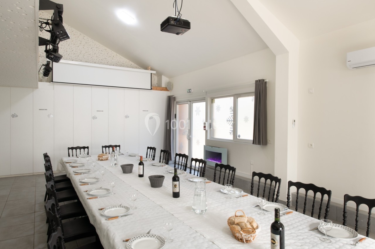 Salle lumineuse avec une grande table dressée pour un repas, équipée de chaises noires et d'un vidéoprojecteur au plafond.