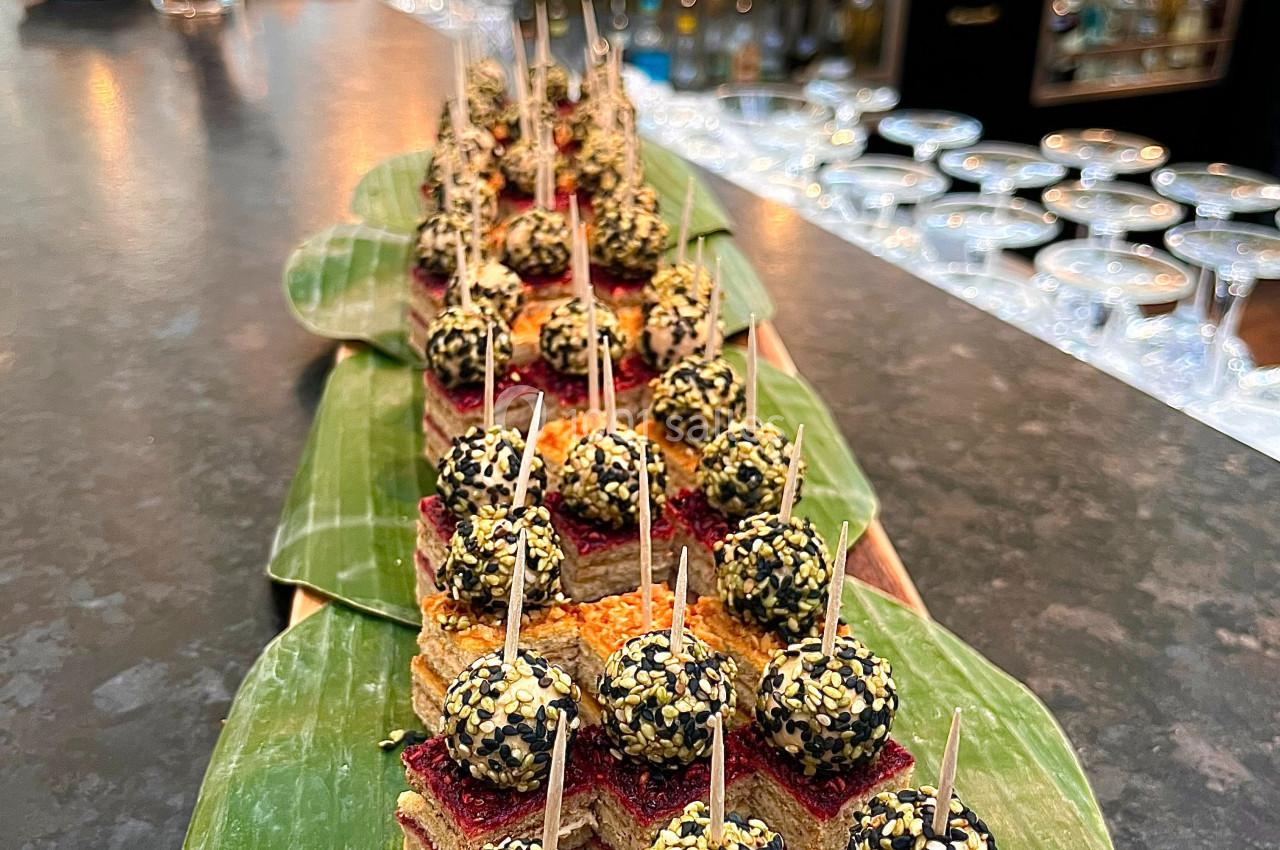 Plateau de bouchées apéritives sur feuilles de bananier, avec des invités en arrière-plan dans un bar animé.