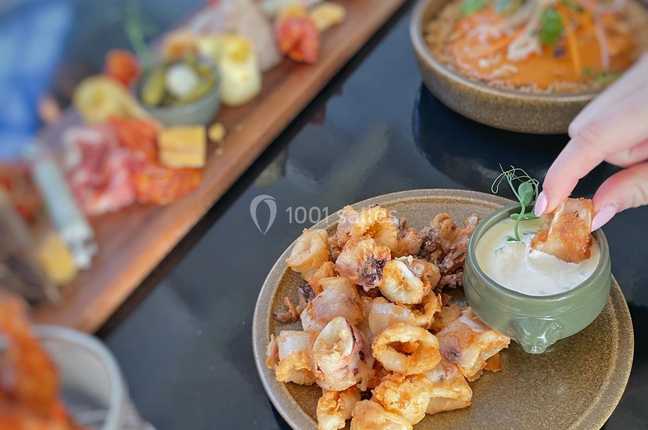 Assiette de calamars frits avec une sauce à tremper, accompagnée de plats variés en arrière-plan sur une table.