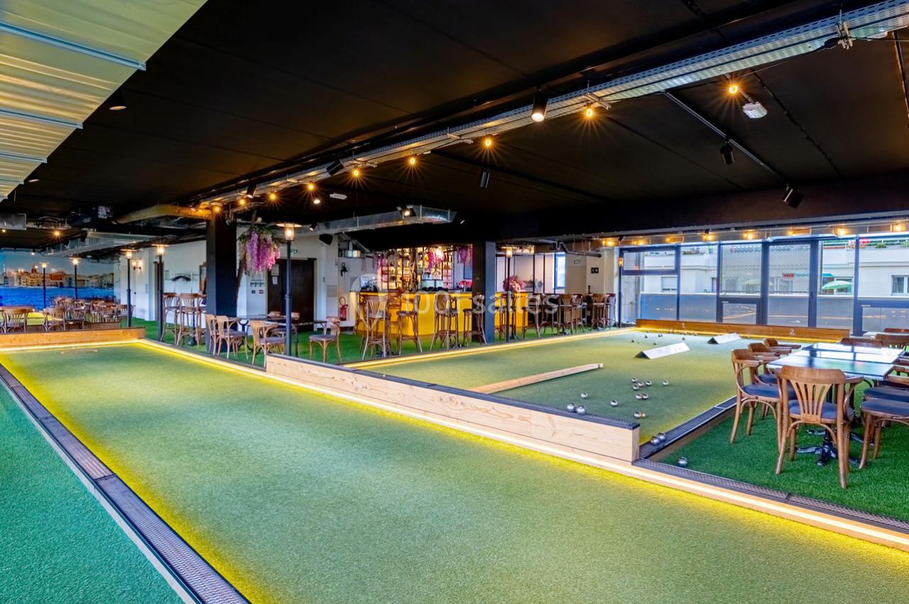 Salle intérieure avec terrain de pétanque en gazon synthétique, tables en bois et éclairage chaleureux.