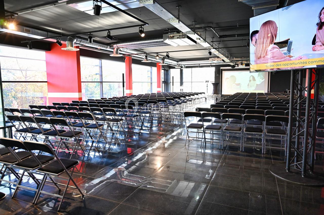Salle de conférence moderne avec des rangées de chaises vides, écran de projection et baies vitrées.