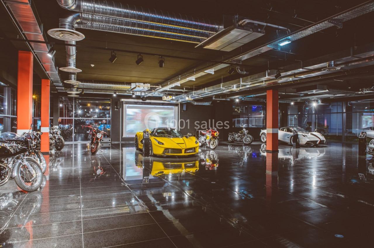 Salle d'exposition moderne avec voitures de sport et motos alignées sur un sol brillant sous un éclairage industriel.