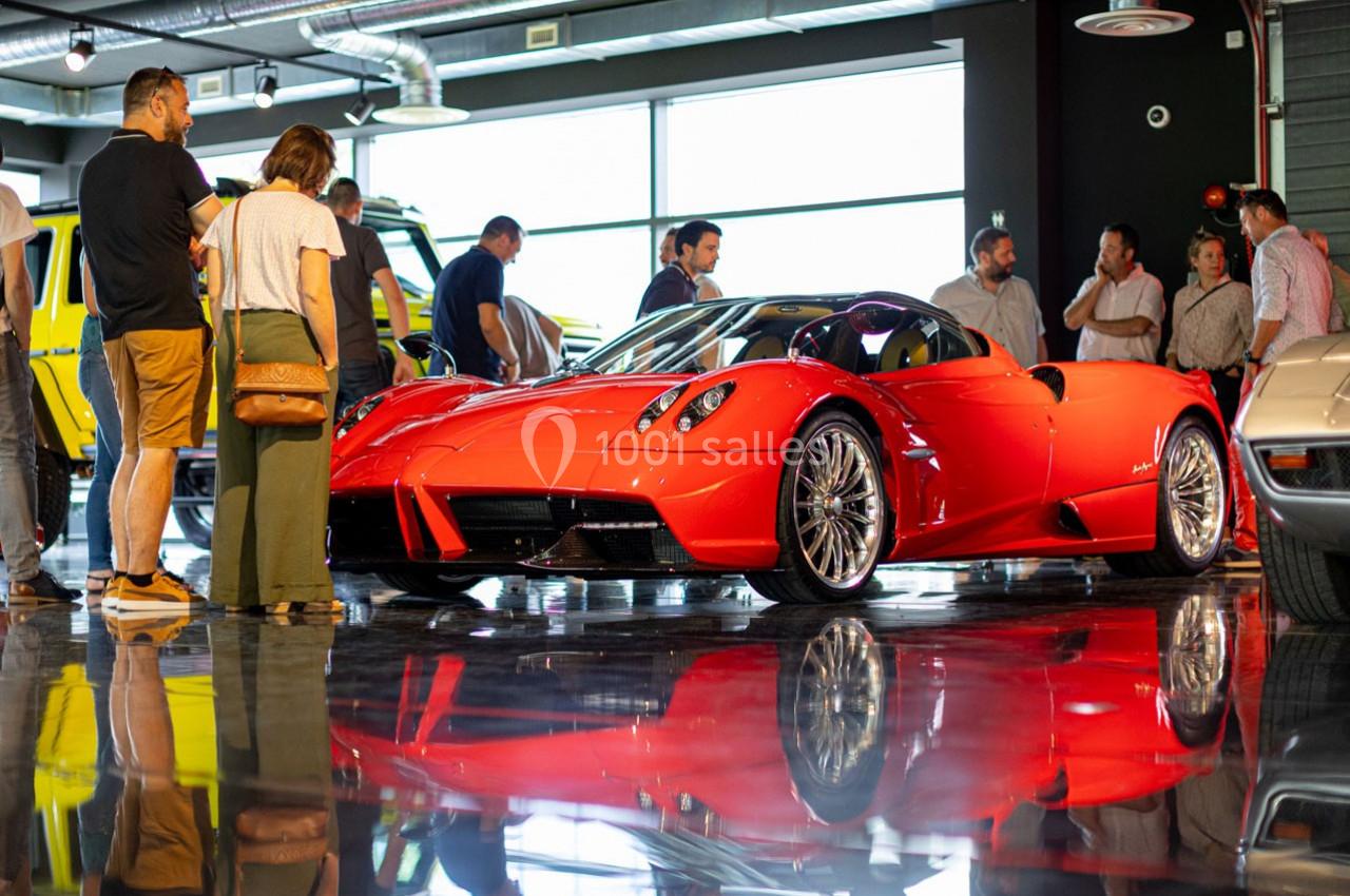 Voiture de sport rouge exposée dans un showroom, entourée de plusieurs personnes en discussion.