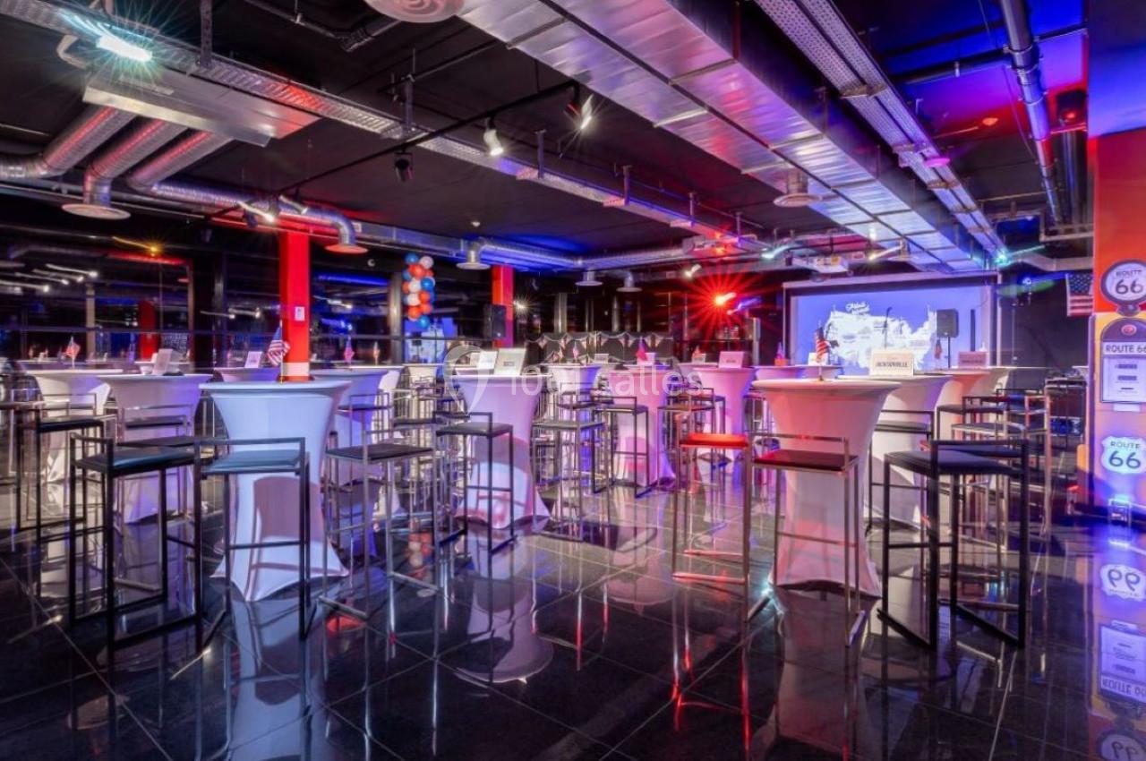 Salle moderne avec tables hautes, tabourets, éclairage coloré et écran de projection en arrière-plan.