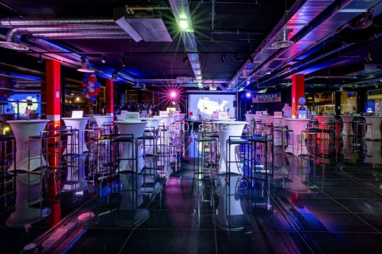 Salle moderne avec tables hautes, tabourets et éclairage coloré, ambiance festive dans un espace intérieur.