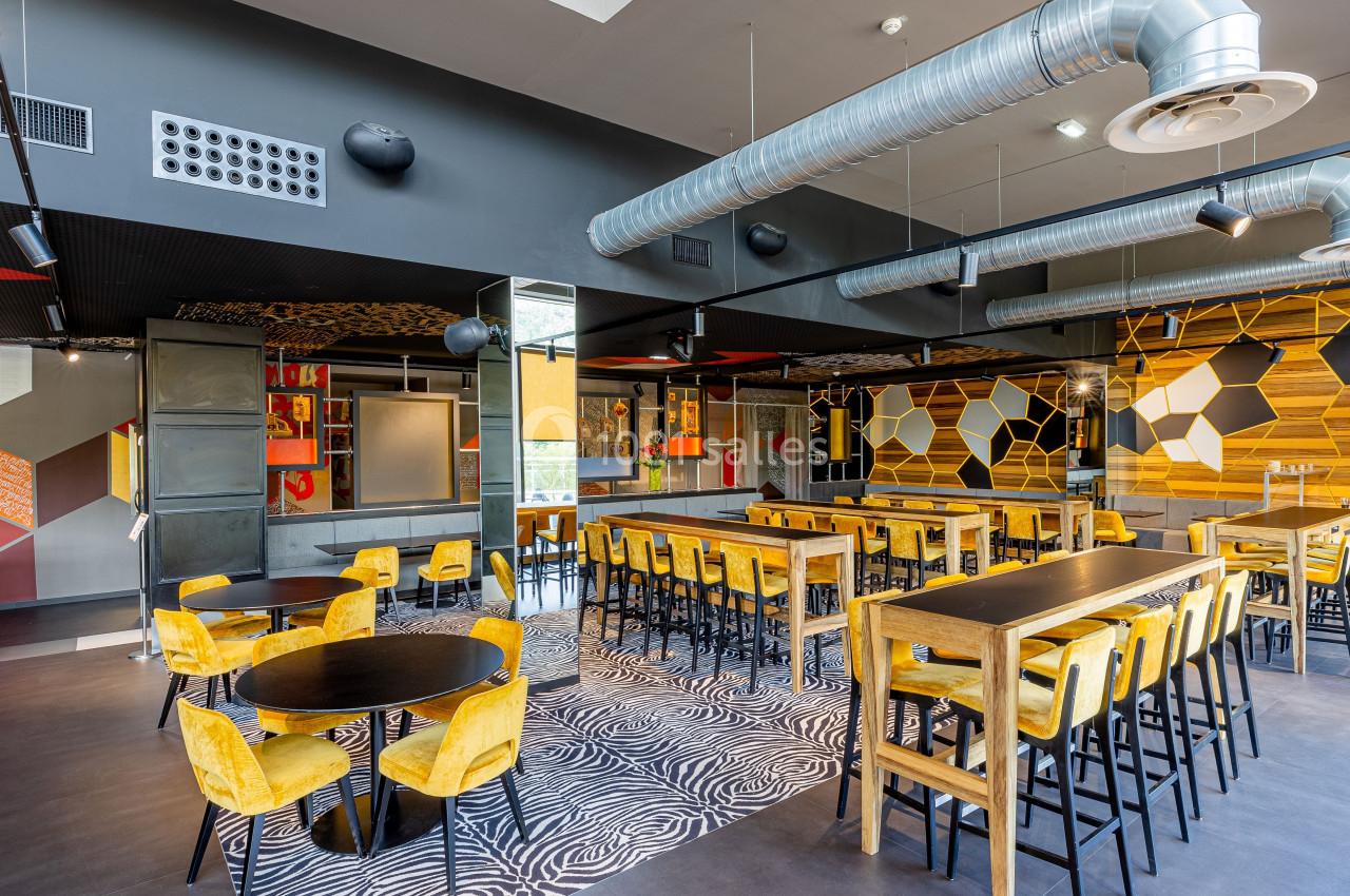 Salle moderne avec mobilier en bois, chaises jaunes, tables hautes et basses, décorations géométriques et conduits apparents.