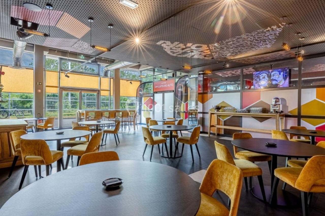 Salle lumineuse avec tables rondes, chaises jaunes, grandes fenêtres et décorations géométriques colorées.