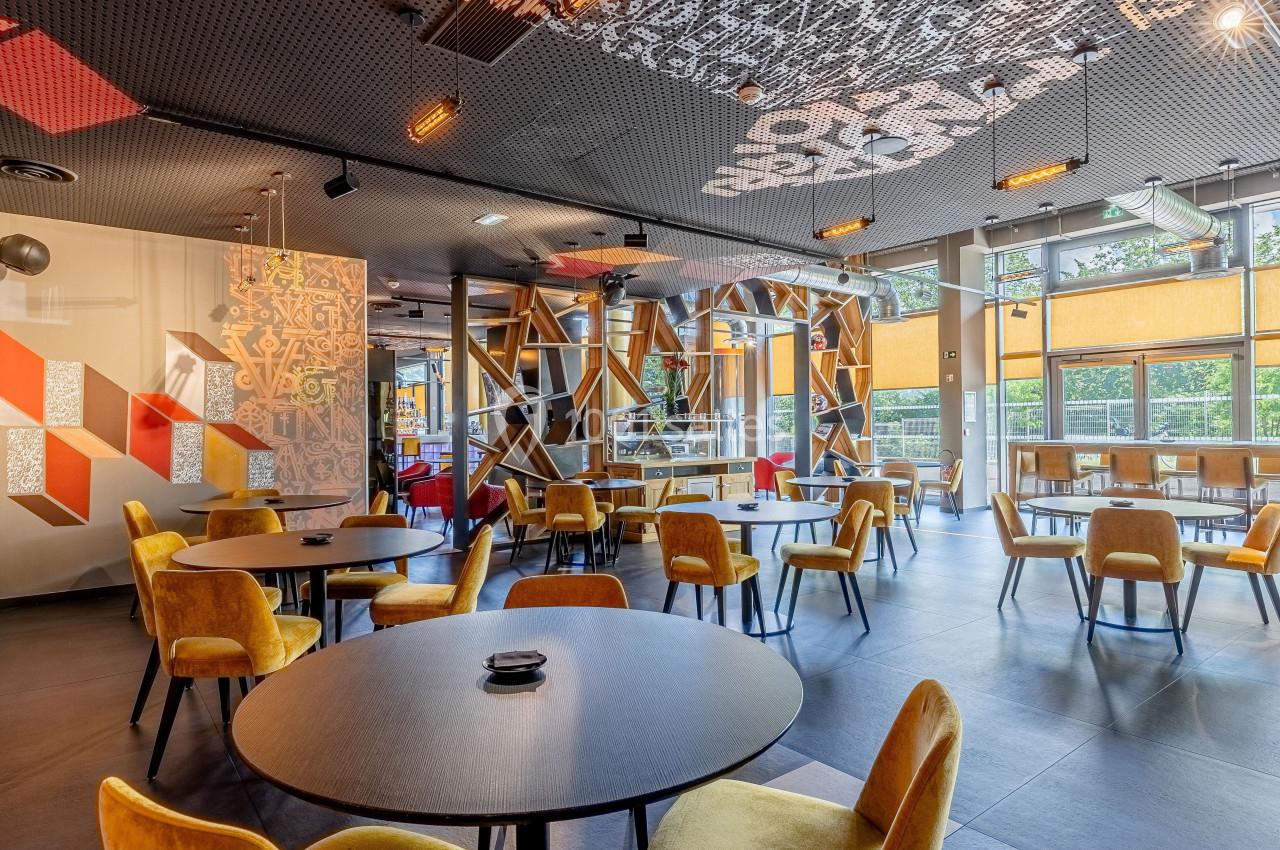 Salle de restaurant moderne avec chaises jaunes, tables rondes noires et grandes baies vitrées laissant entrer la lumière.