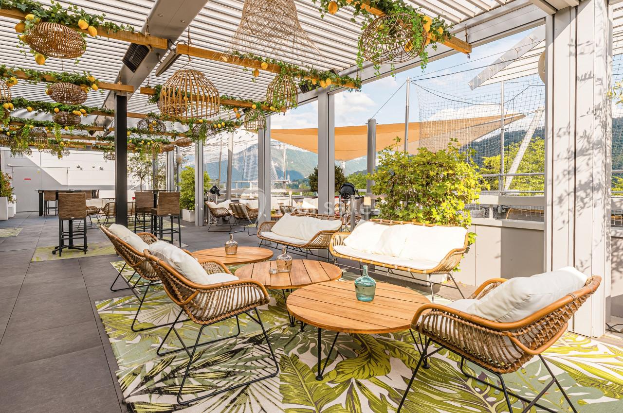 Terrasse aménagée avec mobilier en rotin, tables en bois, plantes suspendues et vue sur des montagnes en arrière-plan.