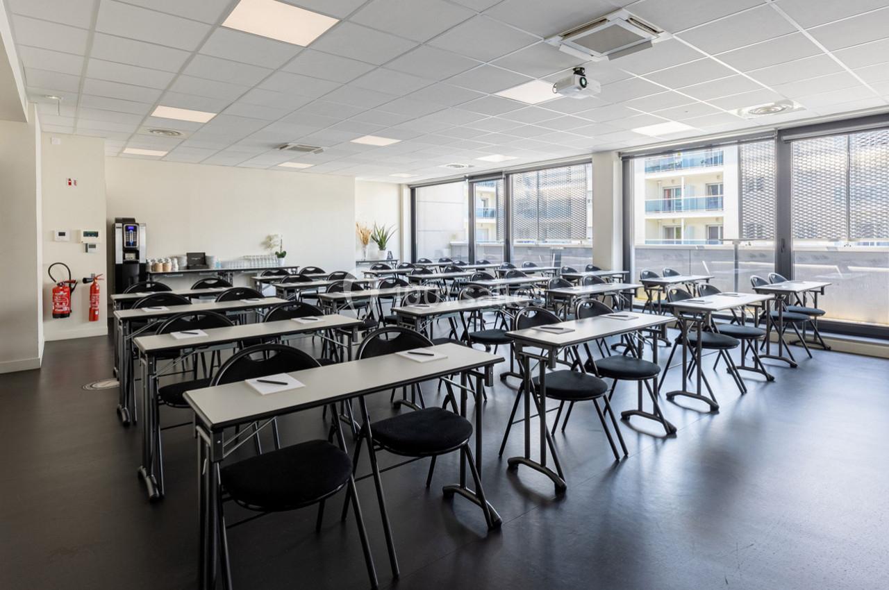 Salle de réunion lumineuse avec des tables et chaises alignées, équipée d'un vidéoprojecteur et d'une machine à café.
