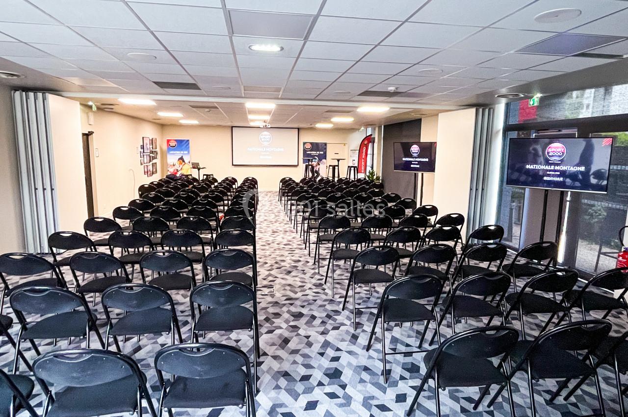 Salle de conférence lumineuse avec rangées de chaises noires alignées face à un écran de projection.