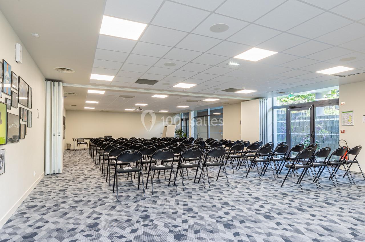 Salle de réunion lumineuse avec des rangées de chaises noires, un sol à motifs et des baies vitrées donnant sur l'extérieur.