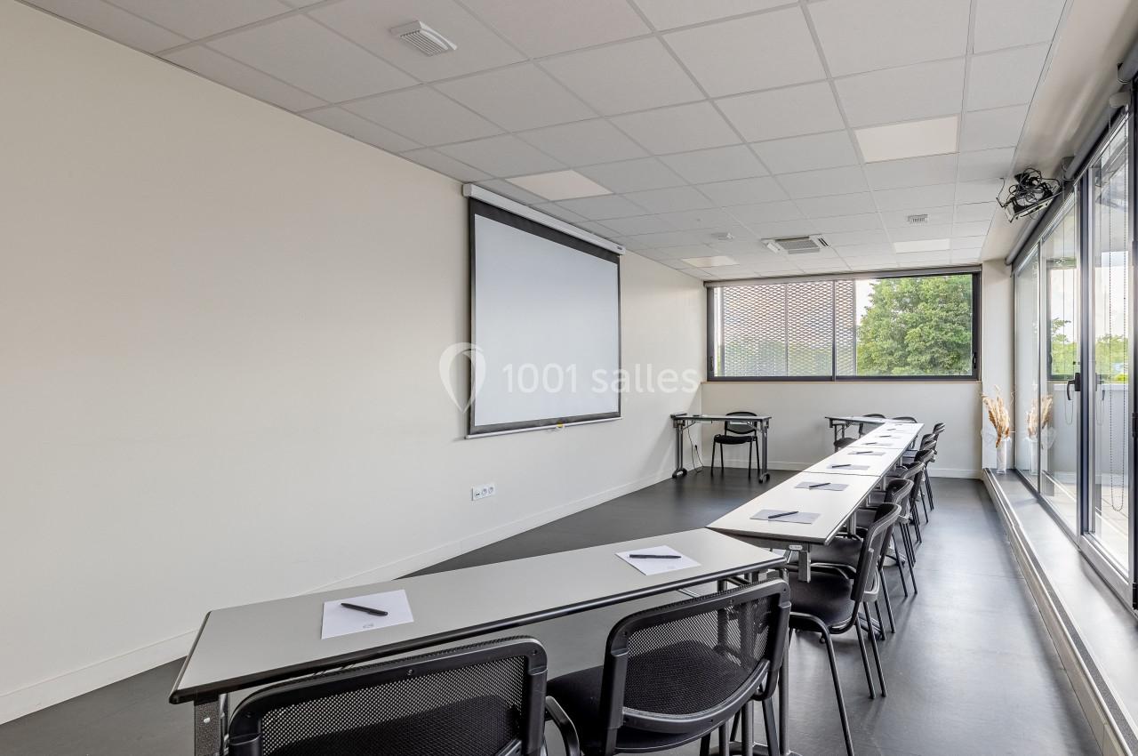 Salle de réunion lumineuse avec tables alignées, chaises noires, écran de projection et grandes fenêtres donnant sur l…