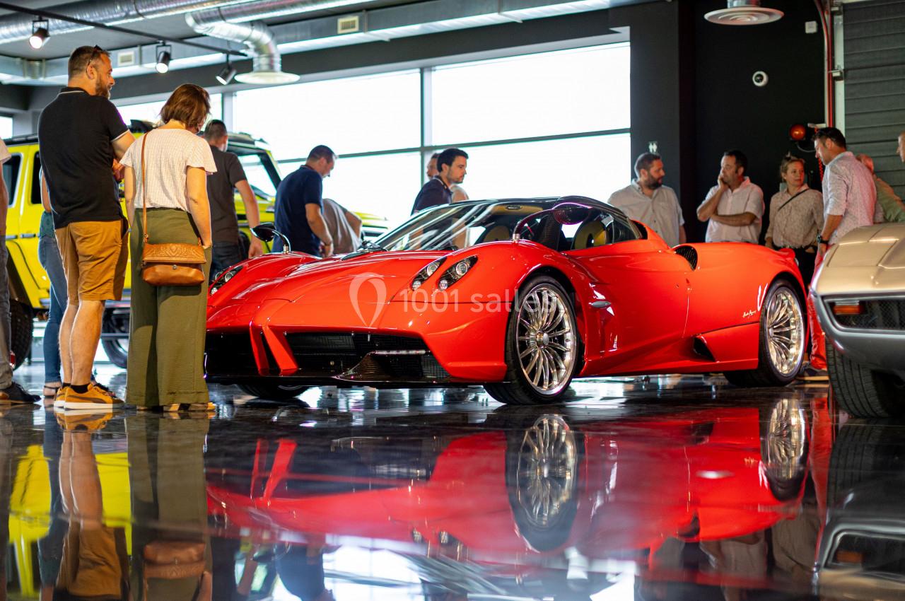 Voiture de sport rouge exposée dans un showroom, entourée de visiteurs et reflétée sur un sol brillant.