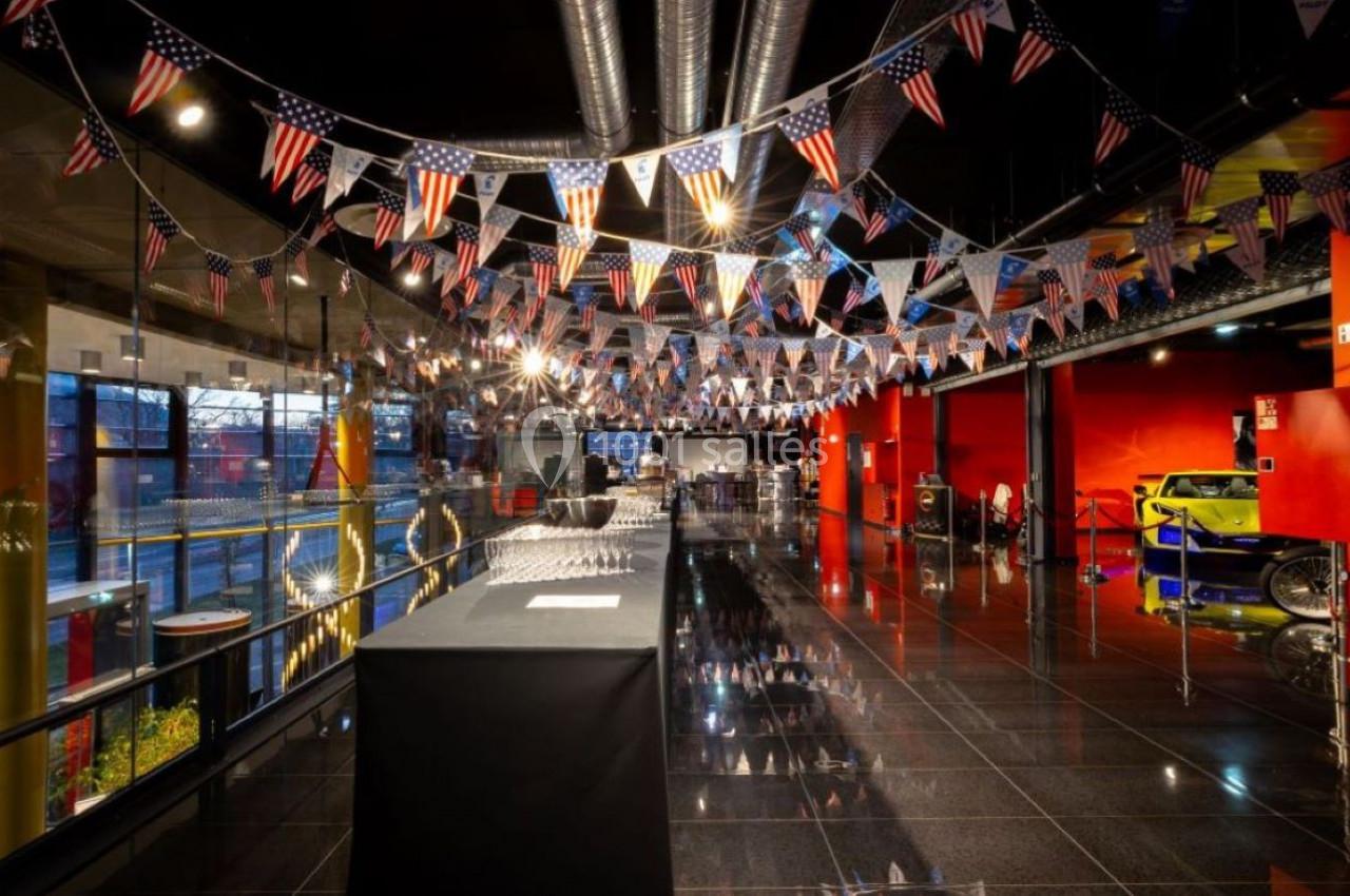 Salle d'exposition décorée de fanions américains, avec des voitures et des éléments industriels en arrière-plan.